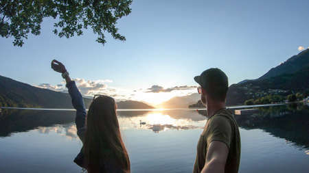 A couple standing at the shore of Millstaetter lake during the sunset. The sun sets behind high Alps. Calm surface of the lake reflects the orange sky and the mountains. They are having fun. Day endの写真素材