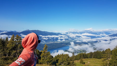 A woman in an orange hoodie enjoying the idyllic view on Alpine Millstaettersee lake in Austria. She...