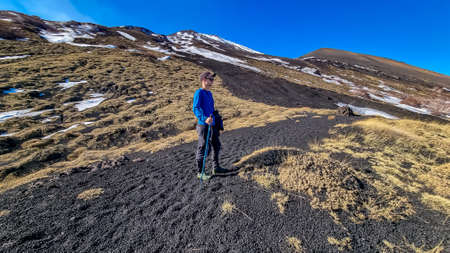 Man surrounded by golden grass hiking on volcanic landscape of volcano mount Etna, in Sicily, Italy, Europe. Barren terrain of solidified lava, ash and pumice. Slopes of crater covered with snowの写真素材