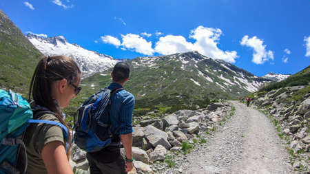 A couple with hiking backpacks hiking in Austrian Alps along a gravelled road. The road is very...