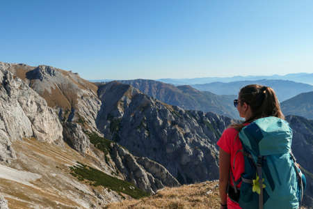 A woman with big blue backpack enjoying the view on valley from top of a mountain in Hochschwab region in Austrian Alps. The flora overgrowing the slopes is turning golden. Freedom. She is happyの写真素材