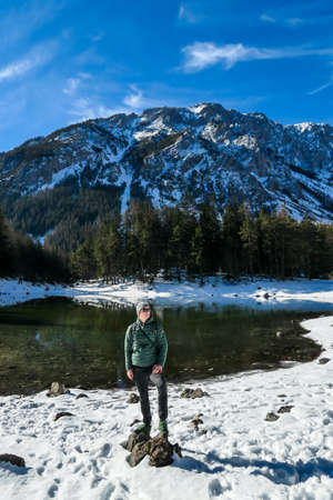 A man standing at the stone next to the shore of Green Lake in Austrian Alps. Mountains and ground are covered with snow. Winter hiking. The man is enjoying a frosty winter day. Winter wonderlandの写真素材