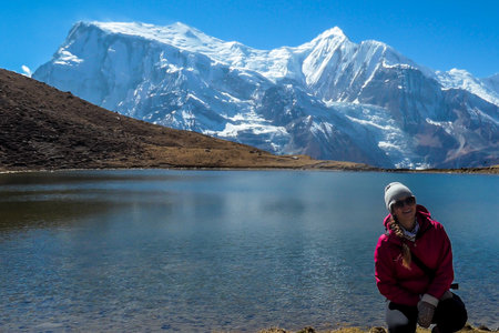 A woman in hiking outfit squatting at the shore of Ice Lake, Annapurna Circuit Trek, Himalayas, Nepal. She is enjoying the idyllic landscape. High, snow caped Annapurna chain in the back. Achievementの写真素材