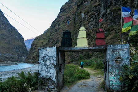 A girl in a hiking outfit crossing through a gate leading to a small Himalayan village along Annapurna Circuit Trek. There is river lowing along the trail. Calmness and peace. Prayer flags on the gateの写真素材
