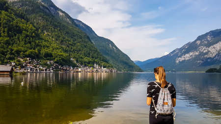 A girl with a small backpack standing at the edge of a lake in Hallstatt, Austria. Girl is looking...