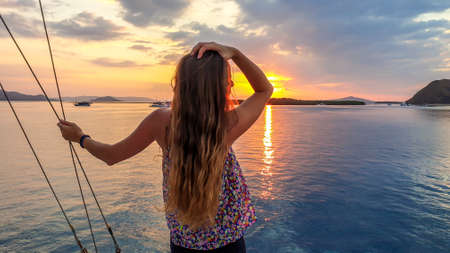 A girl with long hair on a small boat enjoying beautiful sunset in Komodo National Park, Flores, Indonesia. Sun sets over the horizon line. Girl is holding her hair in her hand, trying to tame itの写真素材