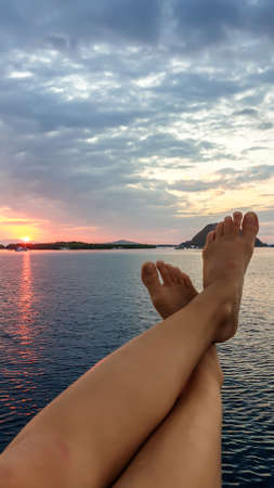 A pair of woman's leg hanging over the rail of small boat, enjoying beautiful sunset in Komodo National Park, Flores, Indonesia. Sun sets over the horizon line. Having a relaxed time.の写真素材