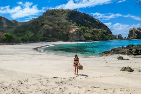 Woman with a straw hat in her hand walking on an idyllic Koka Beach. Hidden gem of Flores Indonesia. She is enjoying solo escape. Waves gently washing the shore. There are hills in the back. Happinessの写真素材