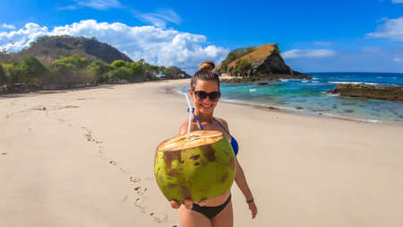 A girl in bikini drinking fresh coconut juice, while sitting on the idyllic Koka Beach and watching the calm sea. Hidden gem of Flores, Indonesia. Island life. Happiness and simplicity. Healthy diet.の写真素材
