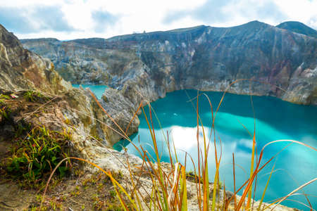 Golden grass growing at the rim of Kelimutu volcanic crater lakes in Flores, Indonesia. Lakes are shining with many shades of turquoise and blue. Sun shines through clouds. Sharp slopes of craterの写真素材