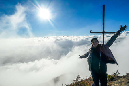 A young woman standing on the top of volcano Inerie in Bajawa, Flores, Indonesia, next to a cross. She is surrounded by thick clouds. Sun tries to get through the thick layer of overcast. Freedomの写真素材
