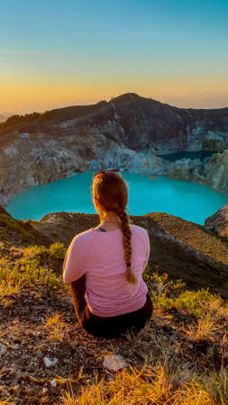 A woman sitting on the ground and watching the sunrise over the Kelimutu volcanic crater lakes in Moni, Flores, Indonesia. Woman is relaxed and calm, enjoying the view on turquoise lake. Golden hourの写真素材