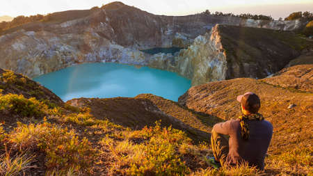 A man watching the sunrise over the Kelimutu volcanic crater lakes in Moni, Flores, Indonesia. Skyline is orange, sun slowly rising. Man is relaxed and calm, enjoying the view on turquoise lakeの写真素材