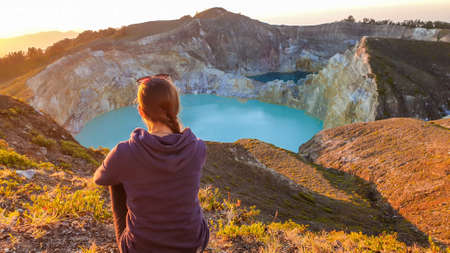 A woman sitting on the ground and watching the sunrise over the Kelimutu volcanic crater lakes in Moni, Flores, Indonesia. Woman is relaxed and calm, enjoying the view on turquoise lake. Golden hourの写真素材