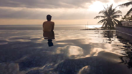 A young man leaning at the edge of an infinity pool and looking at the sun setting into the sea. There is a big palm tree in front. Calmness and serenity. Luxury hotel in Lombok, Indonesia.の写真素材