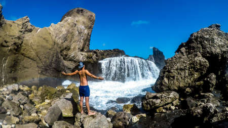 Young man standing in front of the Wave waterfall on Pantai Nambung, Lombok, Indonesia.The water crushes on the rocks on shore, breaching through them, creating a temporary waterfall. Great adventureの写真素材