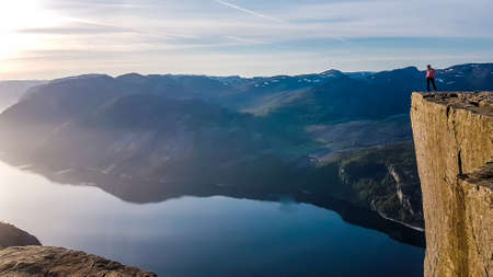 A girl wearing orange hoodie stands at the edge of a steep cliff of Preikestolen during the sunset. A view on Lysefjorden. Fjord goes far inland. Girl enjoys the view, feeling free and happy.の写真素材