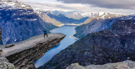 A man wearing blue jacket stands at the hanging rock formation, Trolltunga with a view on...