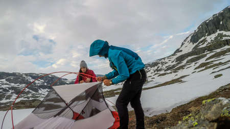 A young couple putting the tent up in the wilderness. The ground is covered with snow. They struggle...