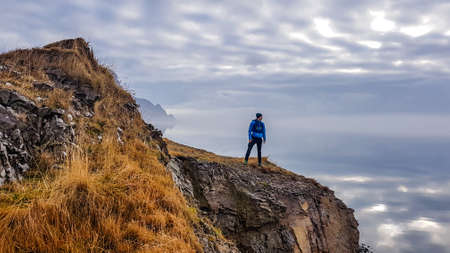 A young man wearing blue jacket stands at the edge of a cliff, looking into the sea. Tall mountains...