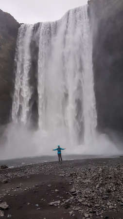 A young man wearing blue jacket standing under the massive waterfall with his arms wide spread. Moody atmosphere. Pure happiness and enjoyment. Gesture of freedom. Beauty of the natureの写真素材