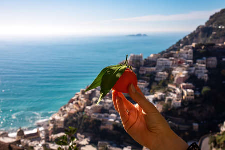Selective soft focus on person holding orange fruit with leaf. Panoramic blurred view on town Positano at Amalfi Coast, Campania, Italy, Europe. View from terrace of luxury hotel at Mediterranean Seaの写真素材