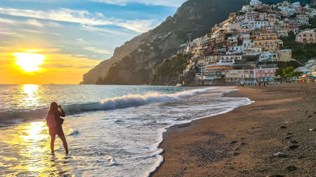 Woman standing in water and watching sunset on Marina Grande Beach and colorful buildings of hillside village Positano, Amalfi Coast, Italy, Campania, Europe. Vacation at Tyrrhenian, Mediterranean Seaの写真素材