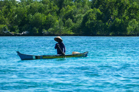Flores/Indonesia 20290808: A fisherman's boat crossing a calm sea near Maumere, Indonesia There is one man in the boat, wearing straw hat and paddling. There is a small green island in the back.のeditorial素材