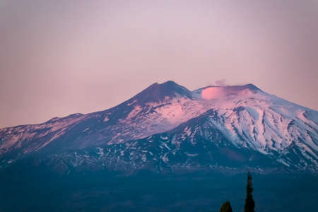 Panoramic view of snow capped Mount Etna volcano during sunrise seen from Taormina, island Sicily, Italy, Europe, EU. Slopes of black lava stones. Snow on peak and sky is colored in vibrant red colorsの写真素材