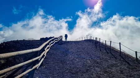 Tourist man hiking along cloudy edge of active volcano crater of Mount Vesuvius, Province of Naples, Campania region, Italy, Europe, EU. Volcanic landscape full of stones, ashes and solidified lavaの写真素材
