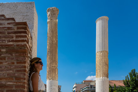 Tourist woman wearing white dress and golden laurel crown leaning against a column of ruined Library...