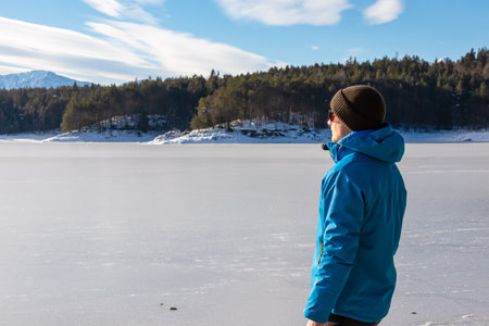 Man in warm hat breathing fresh cold air at frozen alpine lake Forstsee, Techelsberg, Carinthia (Kaernten), Austria, Europe. Winter wonderland at morn. Snow landscape. Karawanks mountain behind forestの写真素材