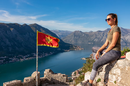 Woman sitting next to Montenegro flag blowing in wind with scenic view from St Ivan fortress on Kotor bay in summer, Adriatic Mediterranean Sea, Montenegro, Balkan, Europe. Fjord along coastal townsの写真素材