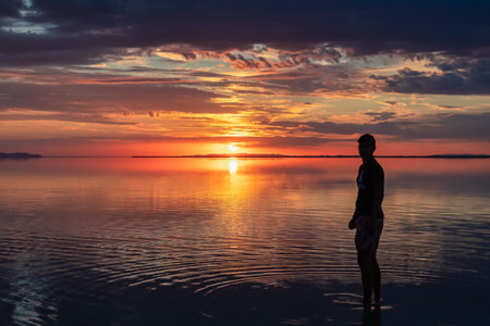 Silhouette of man walking into the sunrise of lake Bonneville Salt Flats, Wendover, Western Utah, USA, America. Dreamy red colored clouds mirroring on the water surface creating romantic atmosphereの写真素材