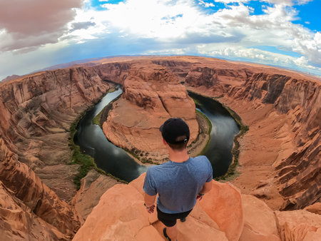 Tourist man standing at the cliff edge with panoramic aerial view of Horseshoe bend on the Colorado river near Page in summer, Arizona, USA. Incised meanders in Glen Canyon National Recreation Areaの写真素材
