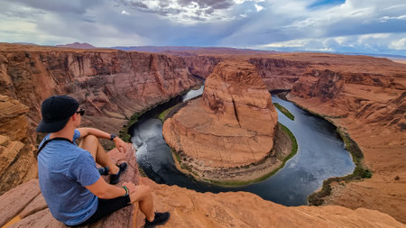 Tourist man sitting at the cliff edge with panoramic aerial view of Horseshoe bend on the Colorado river near Page in summer, Arizona, USA. Incised meanders in Glen Canyon National Recreation Areaの写真素材