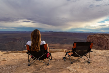 Woman sitting in camping chair enjoying scenic aerial vistas of desert landscape of Valley of the Gods seen from Muley Point near Mexican Hat, San Juan county, Utah, USA. Monument valley in distanceの写真素材