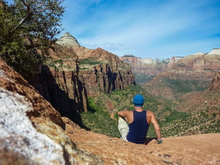 Rear view of man with hat sitting at cliff edge enjoying view of Zion National Park Canyon Overlook, Utah, USA. Adventure seeker embraces nature beauty. Tranquil and majestic atmosphere in wilderness.の写真素材