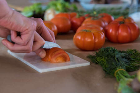 hands holding a knife slicing a tomato with other fresh groceries in backgroundの写真素材
