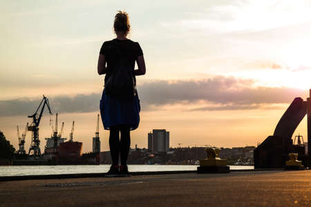 low angle shot silhouette of woman in blue dress standing on pier with harbor structures in backgroundの写真素材