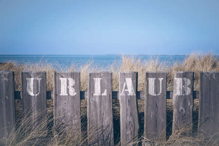 closeup of word Urlaub (German for vacation) written on wooden fence with in front of grass covered dunes and blue sea and sky in the backgroundの写真素材
