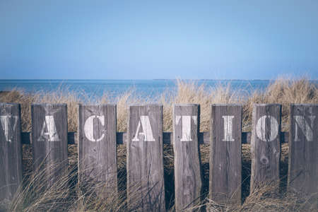 closeup of word vacation written on wooden fence with in front of grass covered dunes and blue sea and sky in the backgroundの写真素材