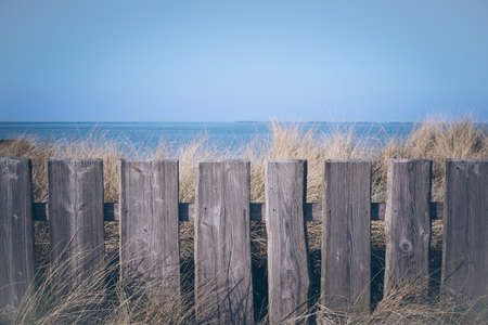closeup of wooden fence in front of grass covered dunes and blue sea and sky in the backgroundの写真素材