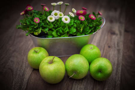 green fresh organic apples on rustic vintage wood table with colorful flowers in metal pot in the backgroundの写真素材
