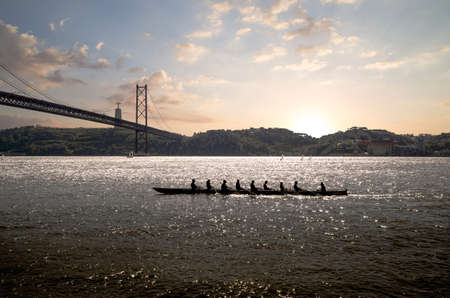 silhouette of people on rowing boat on the sea with suspension bridge in the background at sunset. Lisbon, Portugal.の写真素材