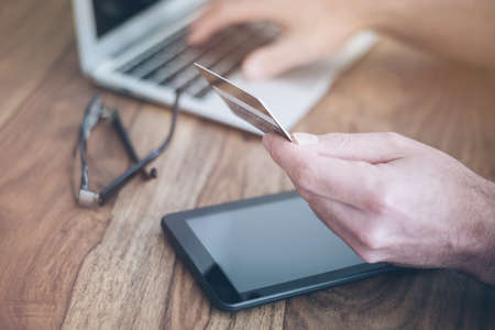 man sitting at rustic wood desk holding a credit card while typing on a laptop computerの写真素材