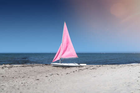 small white sailboat with pink sails on empty beach with ocean and vessels on distant horizon under clear blue skyの写真素材