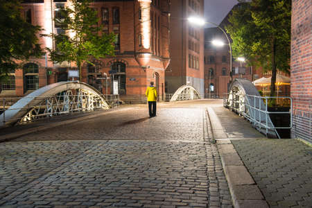 long exposure of young woman in yellow raincoat standing on bridge in illuminated old warehouse district Speicherstadt in Hamburg, Germany at nightの写真素材