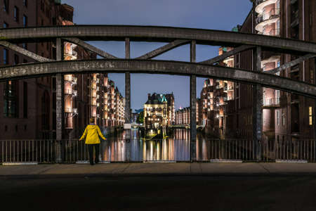 long exposure of young woman in yellow raincoat standing on bridge in illuminated old warehouse district Speicherstadt in Hamburg, Germany at nightの写真素材