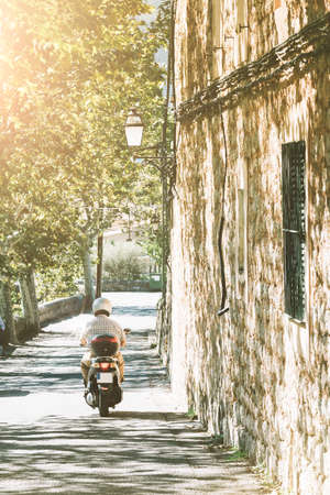 male person on motor scooter driving down narrow street in mediterranean villageの写真素材